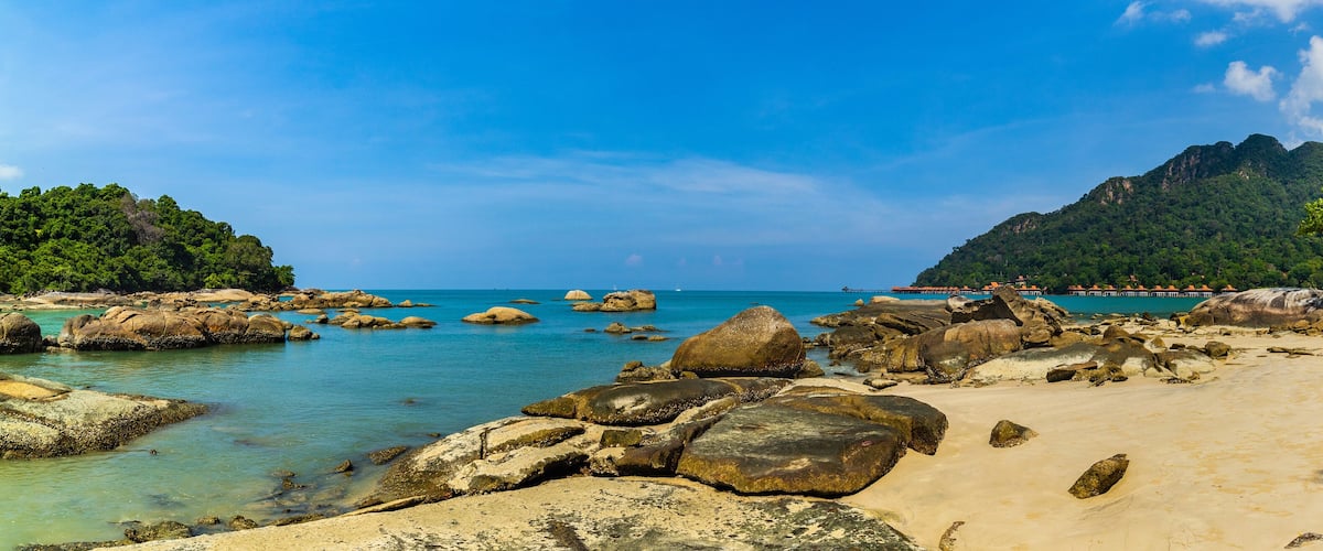 A beach with large rocks, clear turquoise water, and a mountainous backdrop.