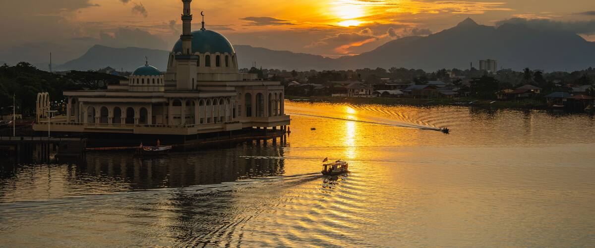 A boat on a calm river with a large building featuring a blue dome and a tall minaret, set against a sunset sky and mountains in the background.
