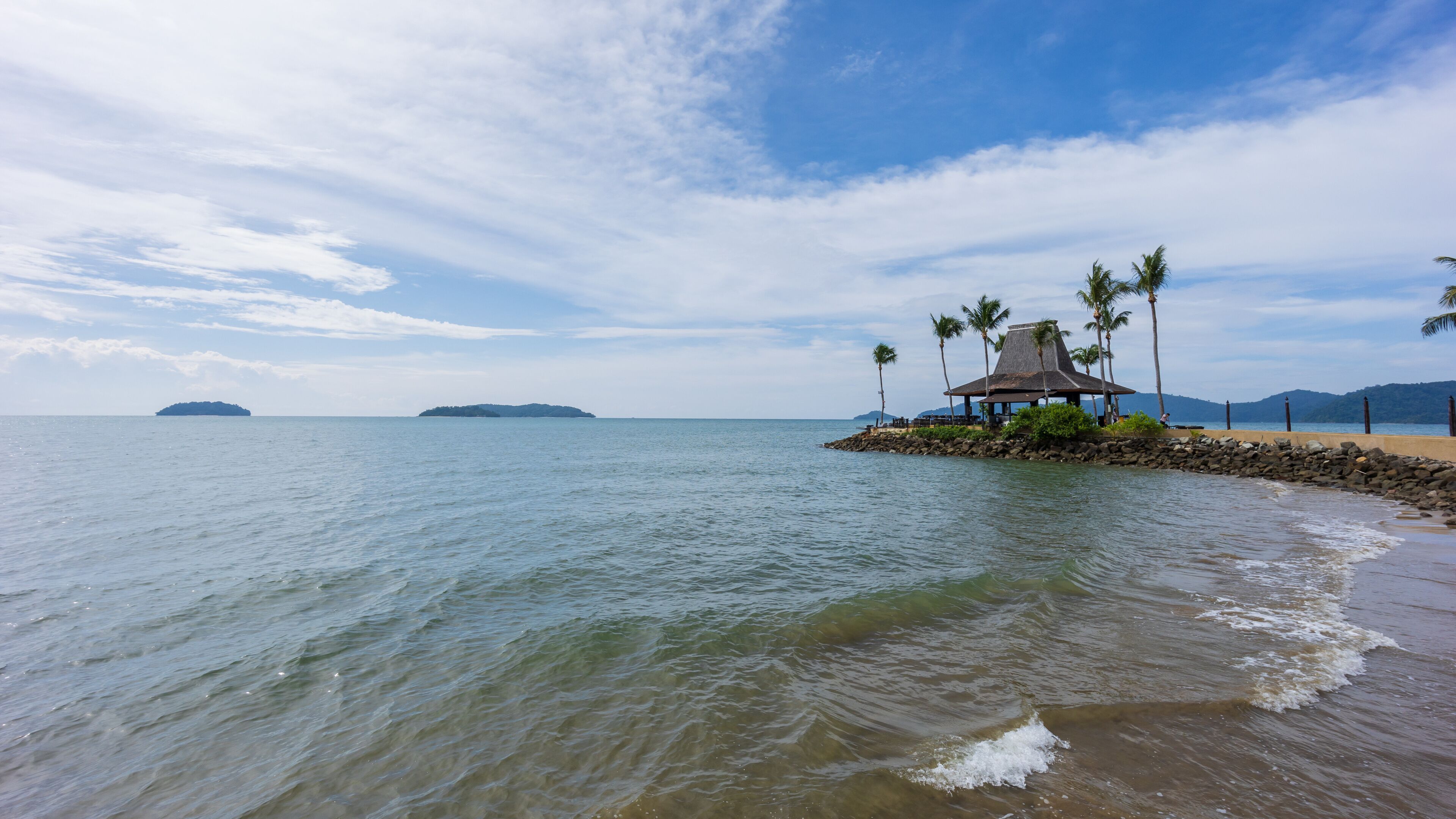 A beach with a thatched-roof hut surrounded by palm trees, calm water, and distant islands.