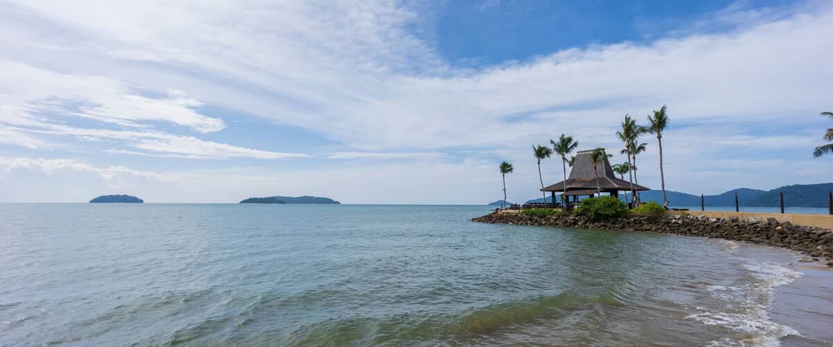 A beach with a thatched-roof hut surrounded by palm trees, calm water, and distant islands.