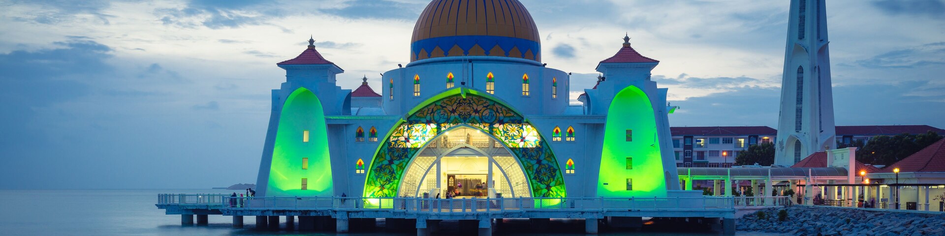 Una mezquita con una cúpula grande y una entrada iluminada en verde, rodeada de agua y un minarete alto.