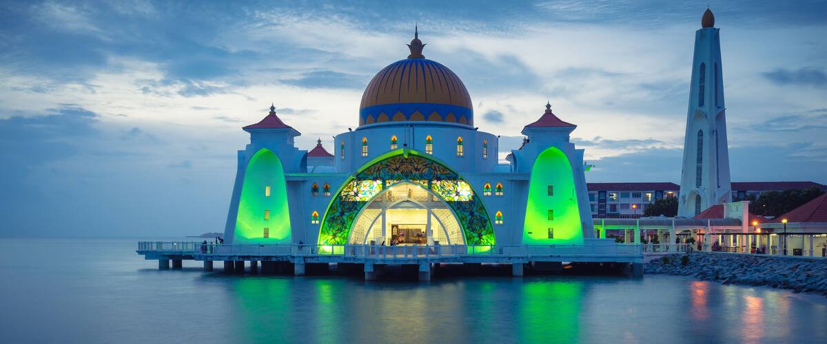 A mosque with a large dome and green-lit entrance, surrounded by water and a tall minaret.