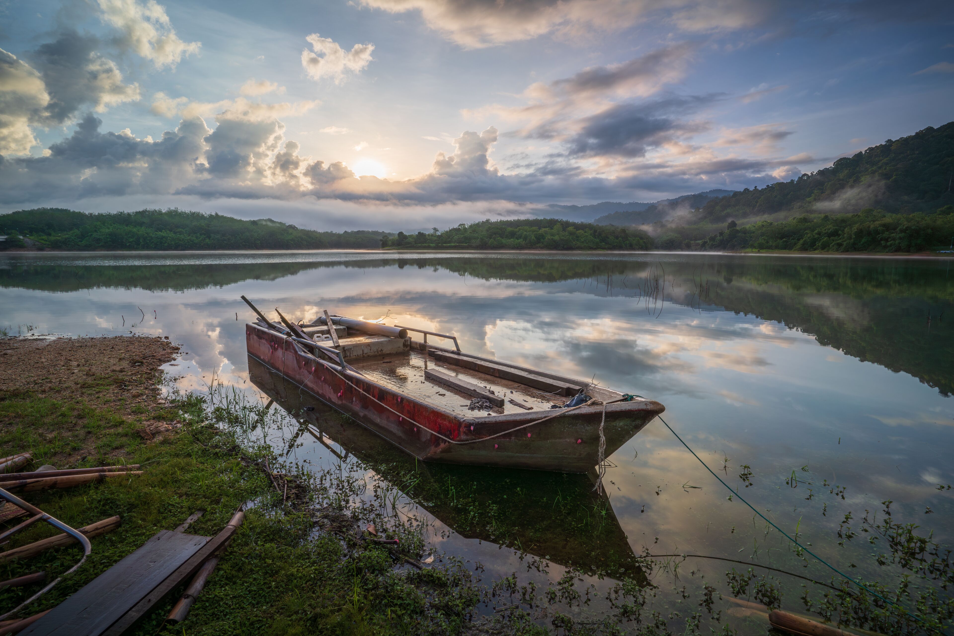A calm lake with a small boat tied to the shore, surrounded by lush greenery and hills.