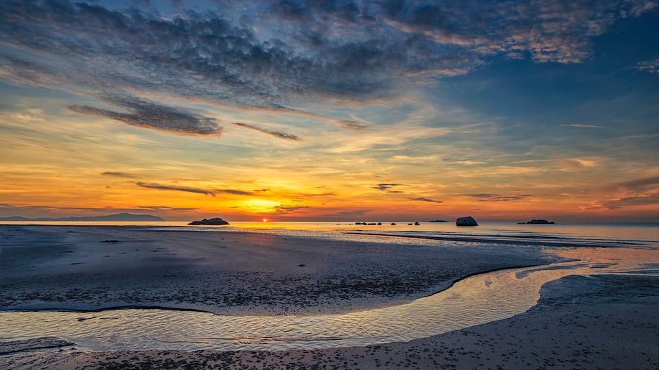 Ein Sonnenuntergang über einem Strand mit ruhiger See und vereinzelten Felsen.
