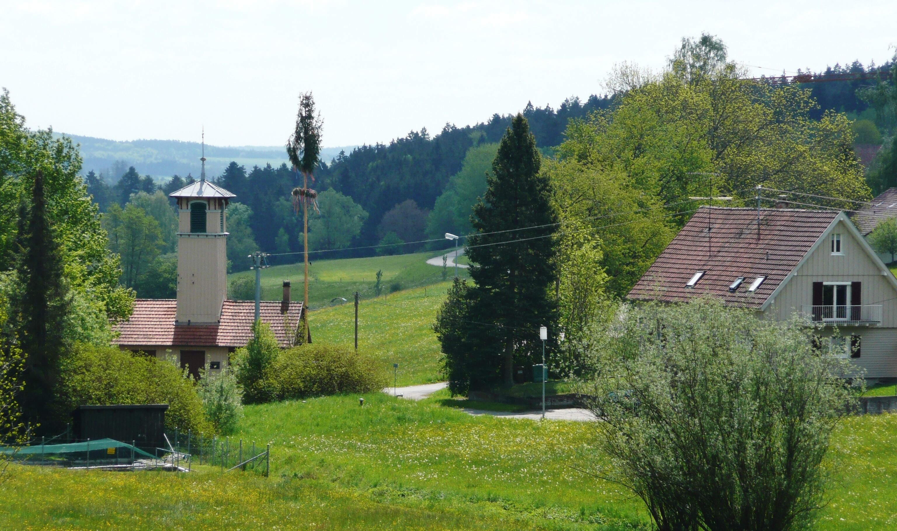 A rural landscape with a church tower, a house with a red roof, and a green field with trees.