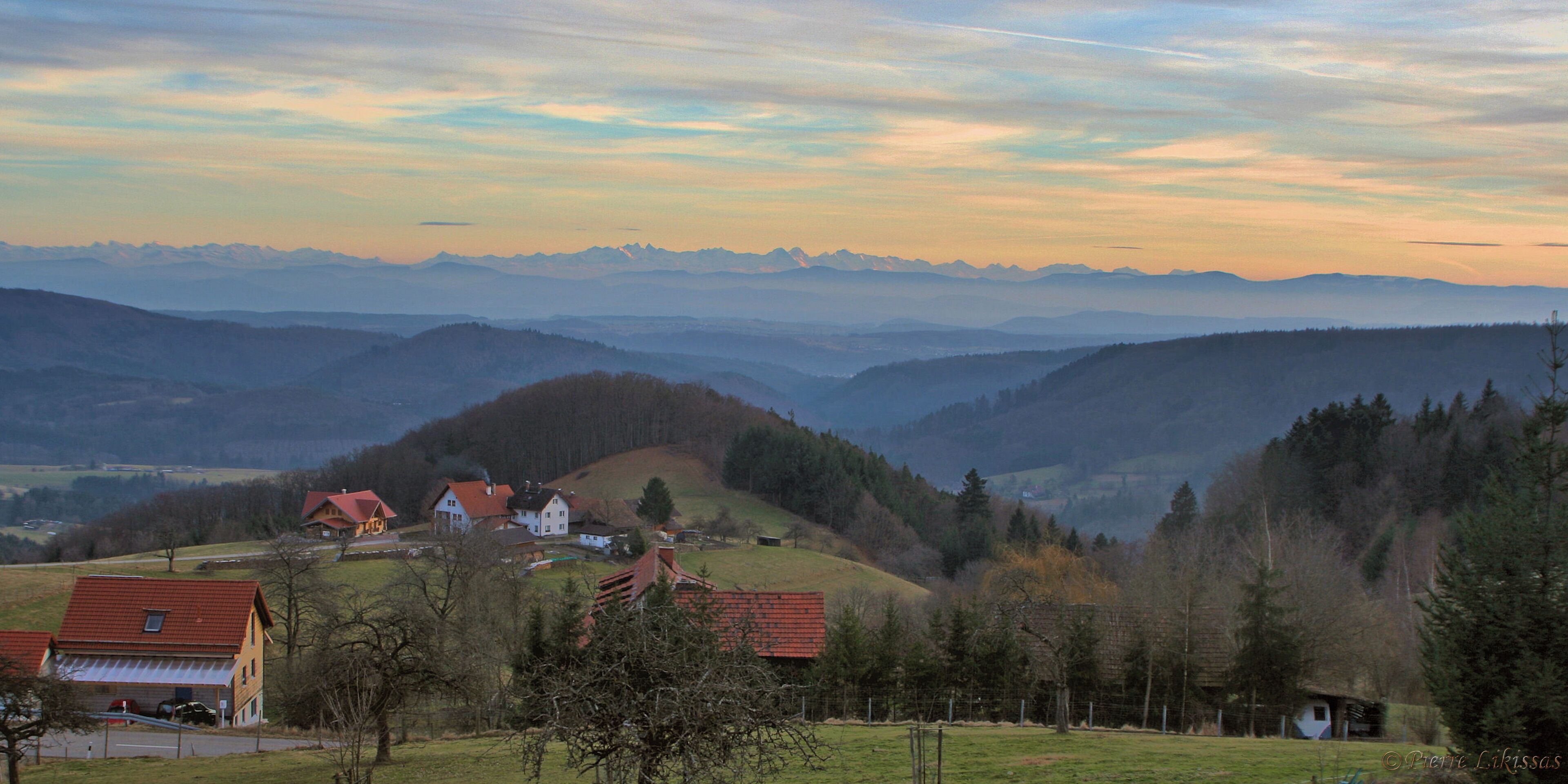 A rural landscape with rolling hills, a small village with red-roofed houses, and distant mountains under a colorful sky.