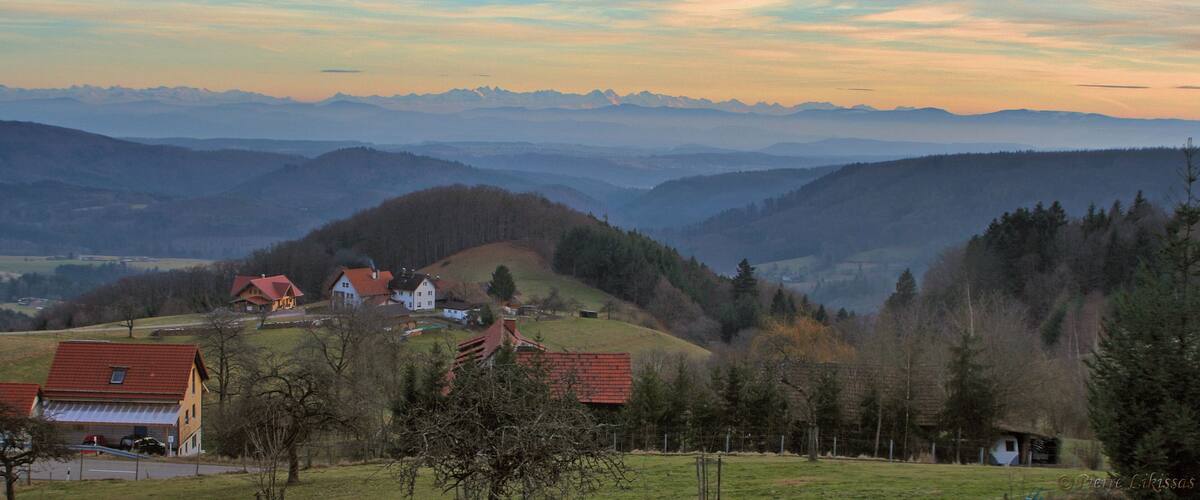 A rural landscape with rolling hills, a small village with red-roofed houses, and distant mountains under a colorful sky.
