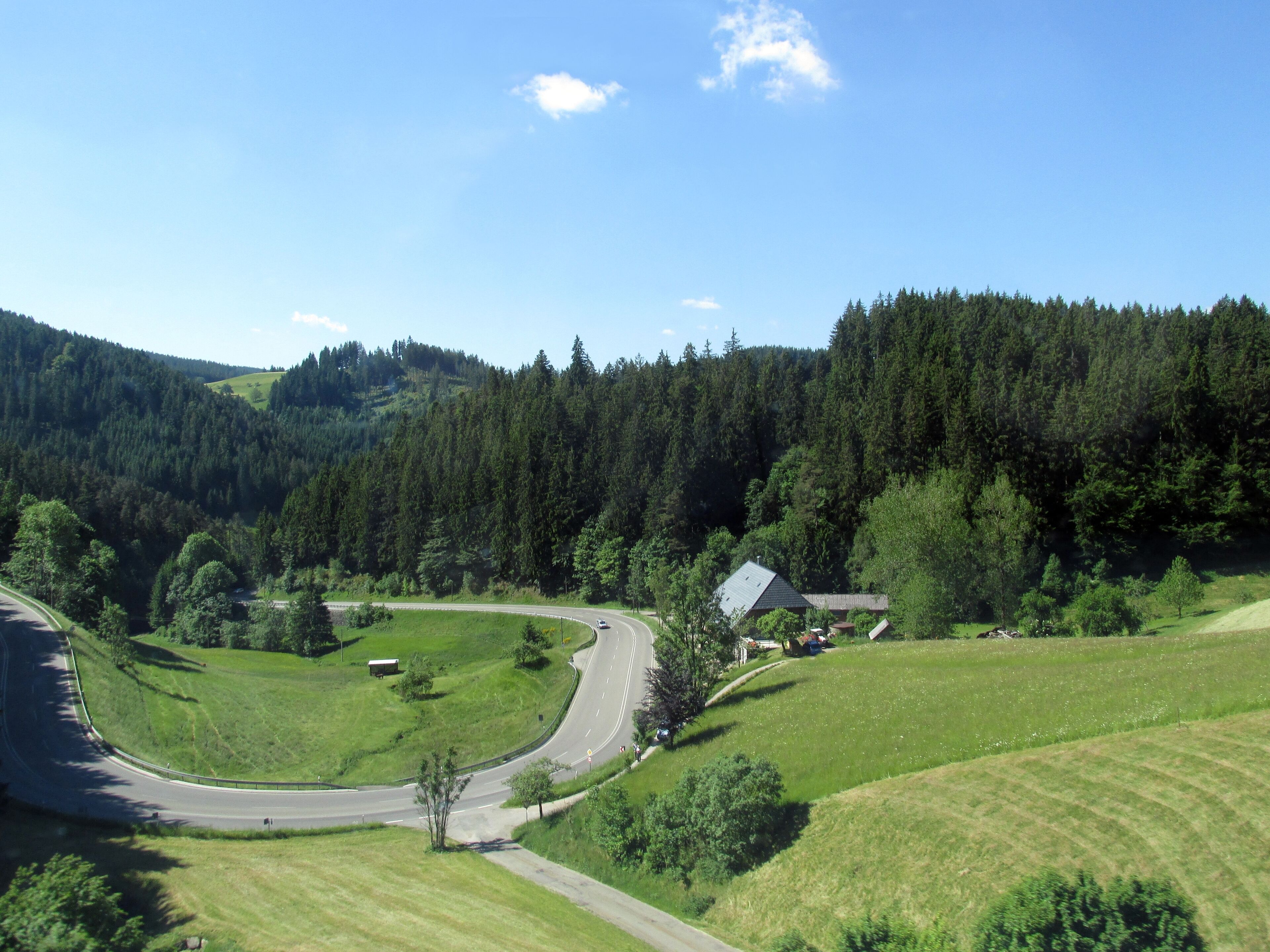 A winding road through a green valley with a house and a forested hill in the background.