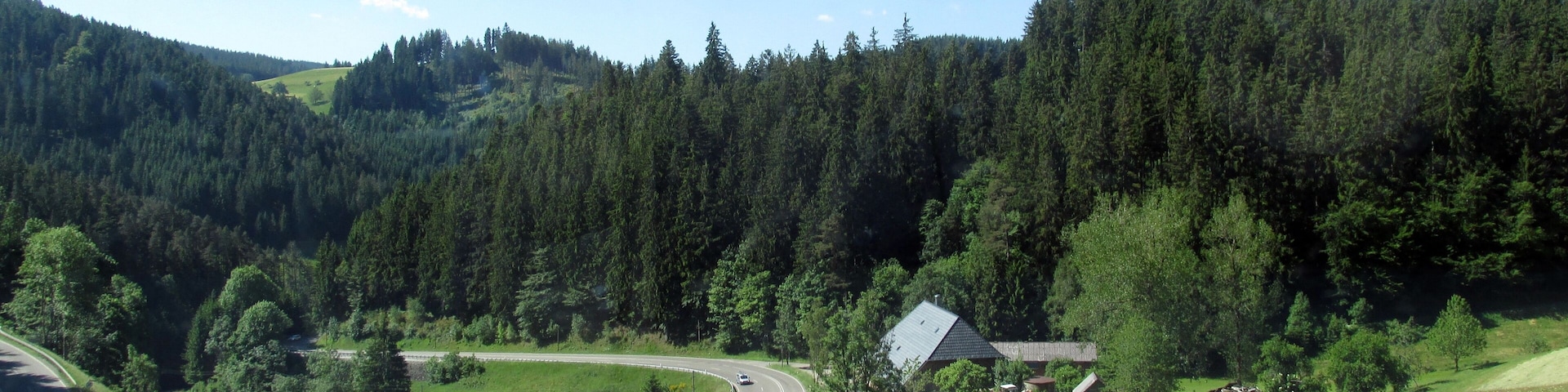 A winding road through a green valley with a house and a forested hill in the background.