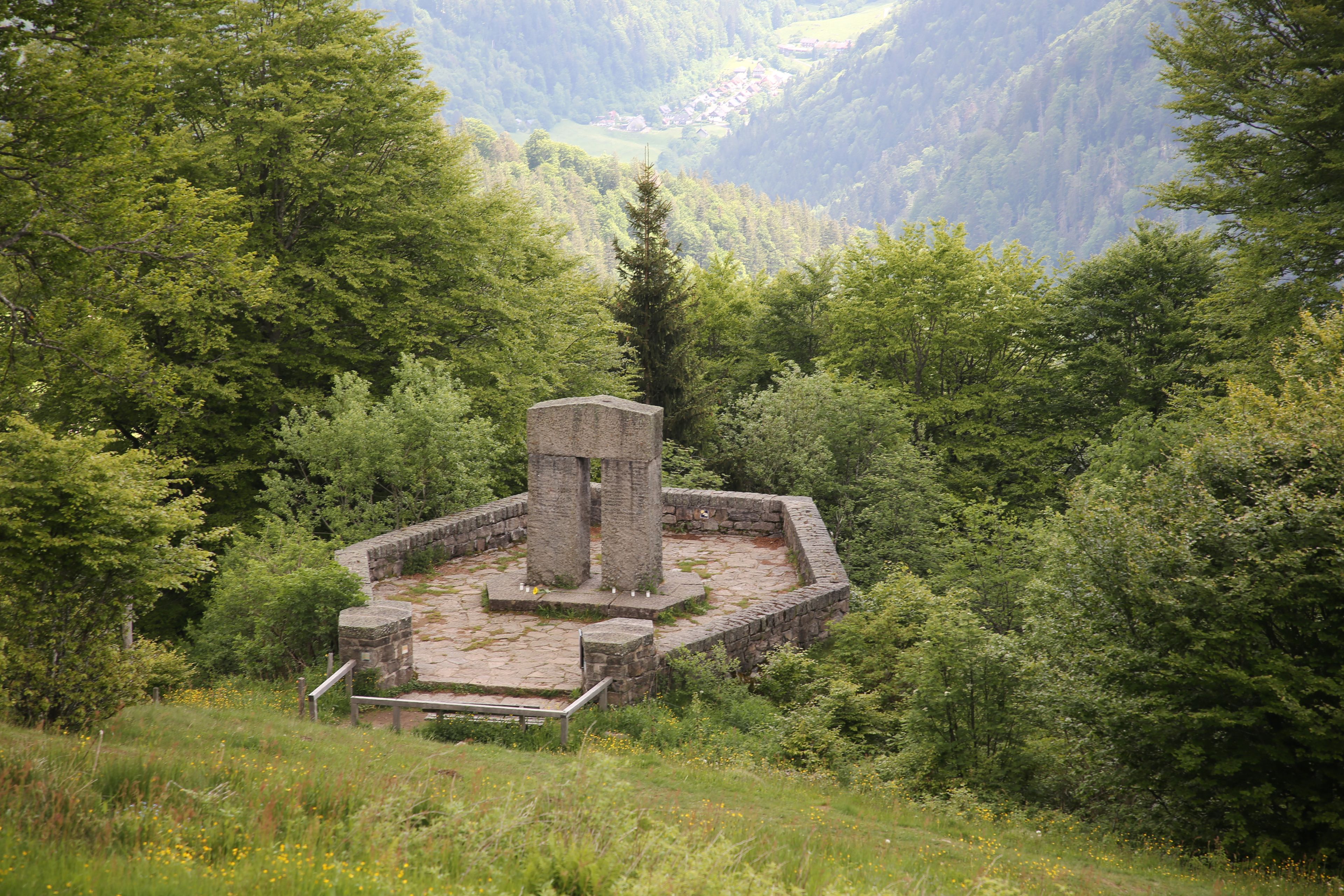 Eine steinerne Ruine mit einer zentralen Säule, umgeben von einer Steinmauer, inmitten eines bewaldeten Gebiets mit Bergen im Hintergrund.