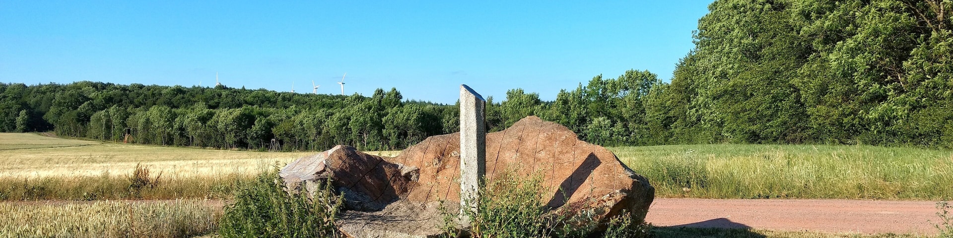 Paisaje rural con una gran formación rocosa, un campo de trigo y un bosque al fondo.