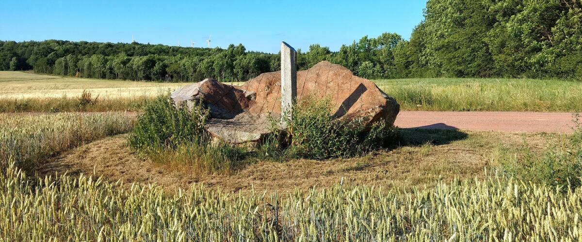 A rural landscape with a large rock formation, a wheat field, and a forest in the background.
