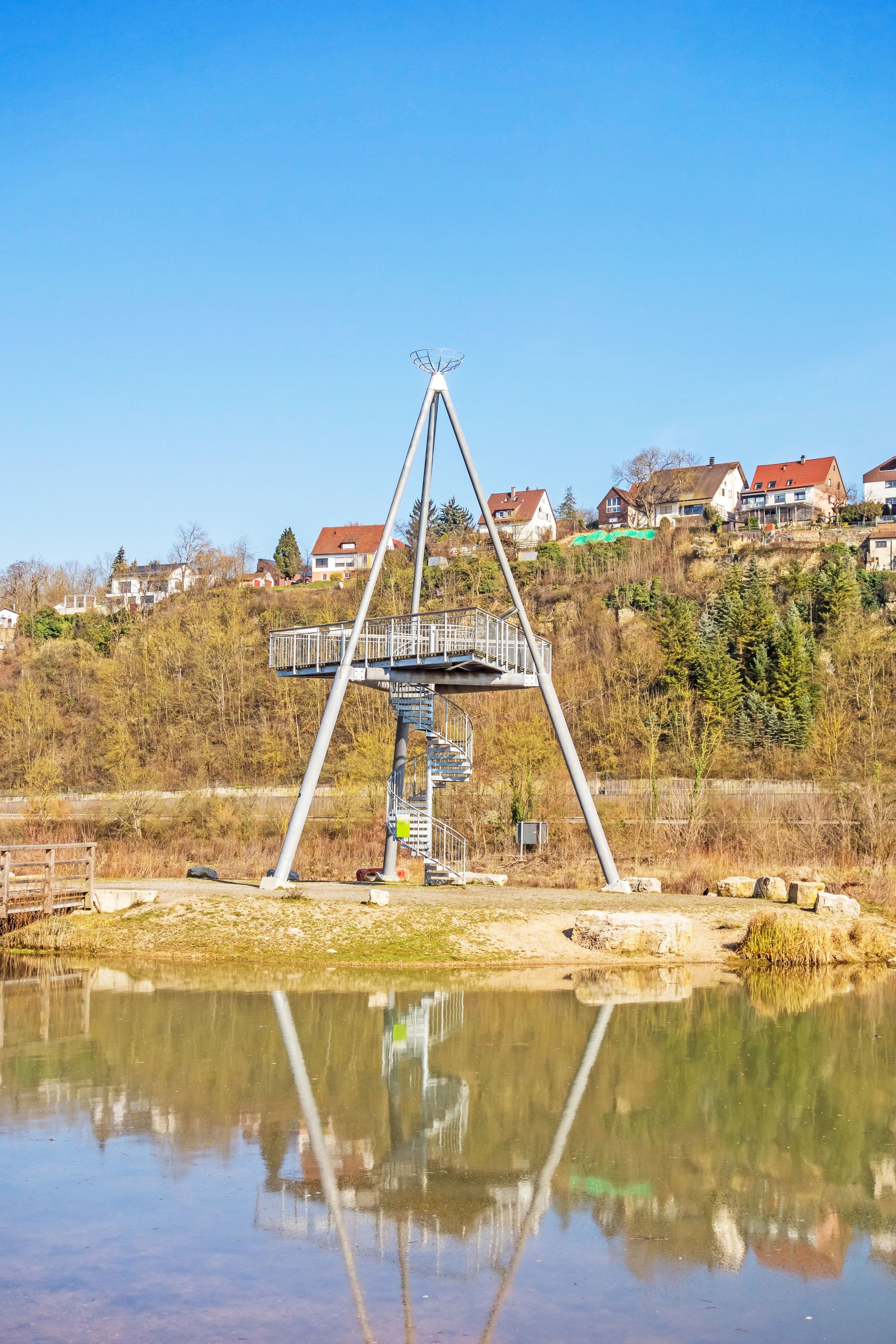 A metal structure with a staircase and a platform, surrounded by a body of water with reflections, set against a backdrop of houses and trees.