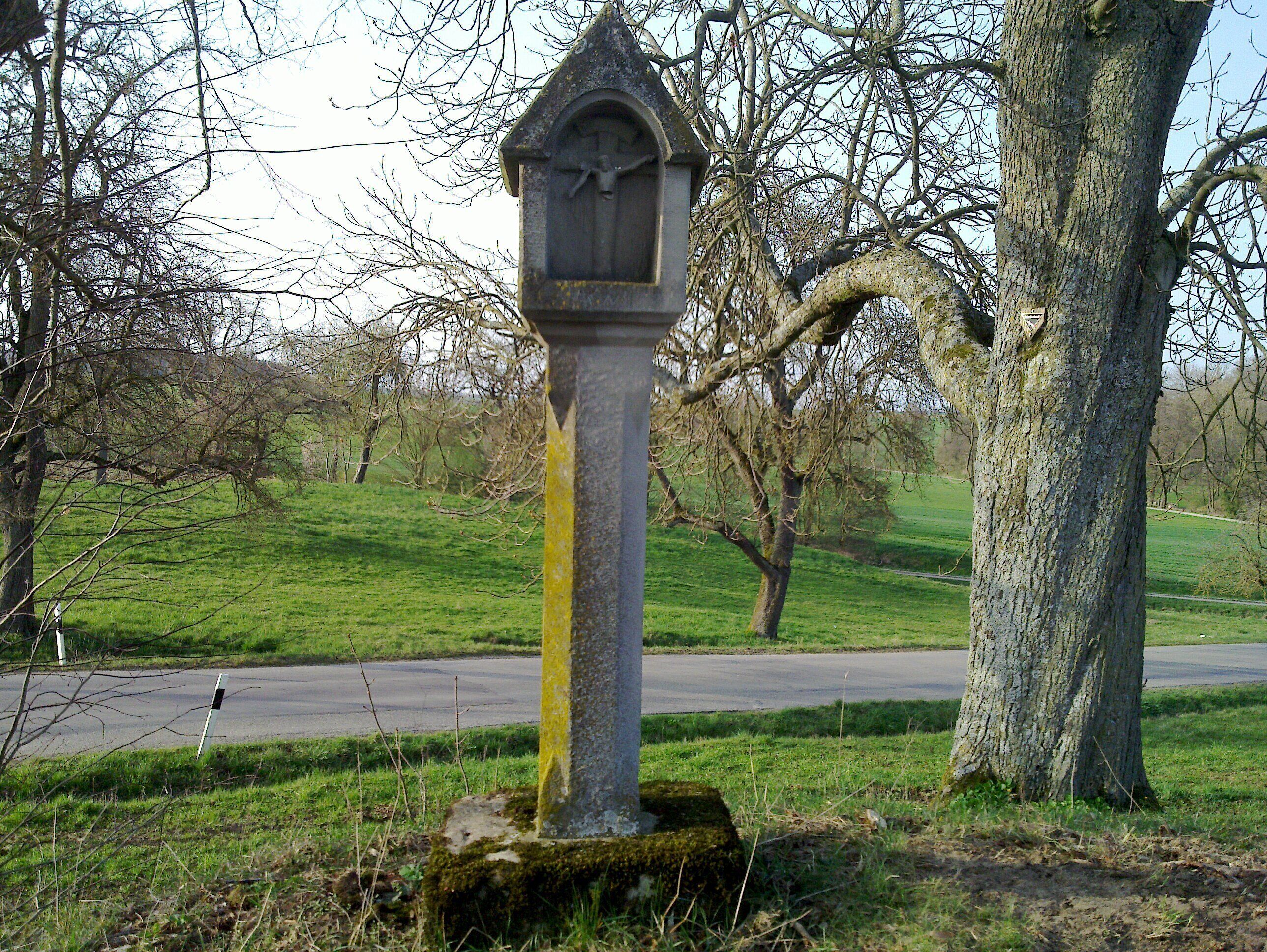 A stone cross with a crucifix, surrounded by grass and trees.
