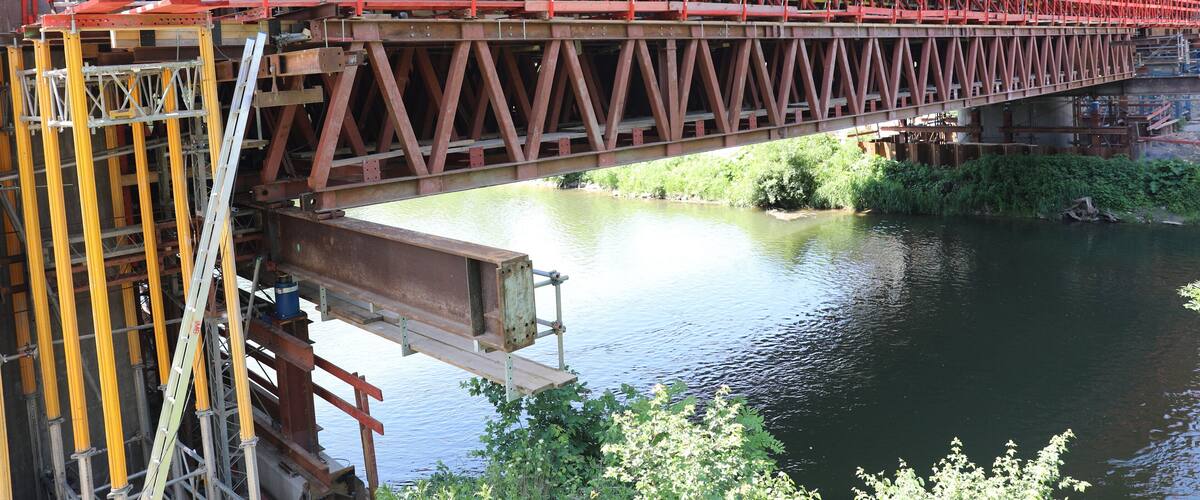 A large red metal bridge spanning a calm river with greenery around.