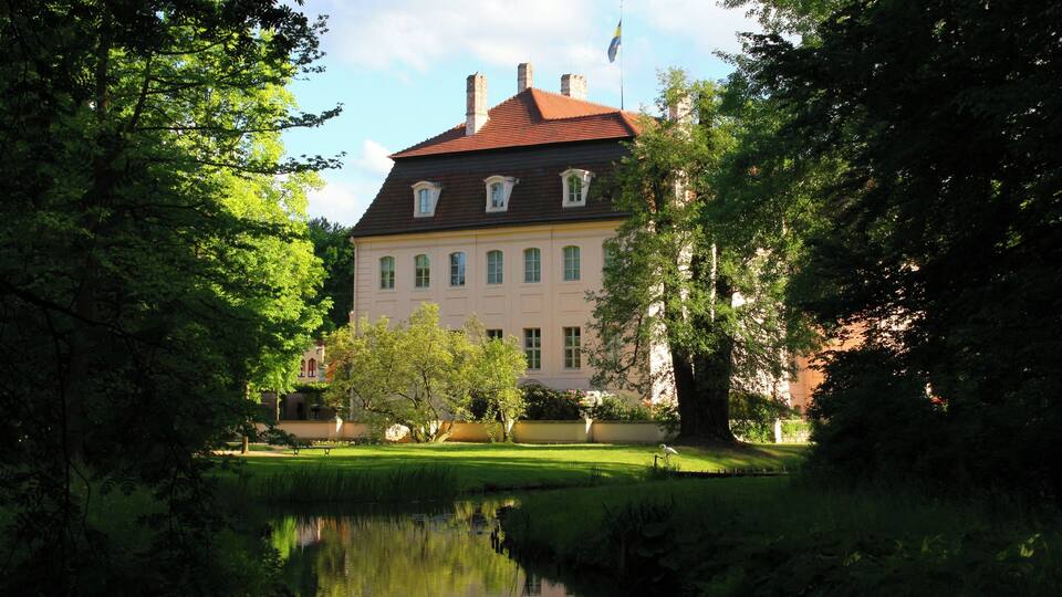 A large building with a red roof, surrounded by lush greenery and a calm pond.