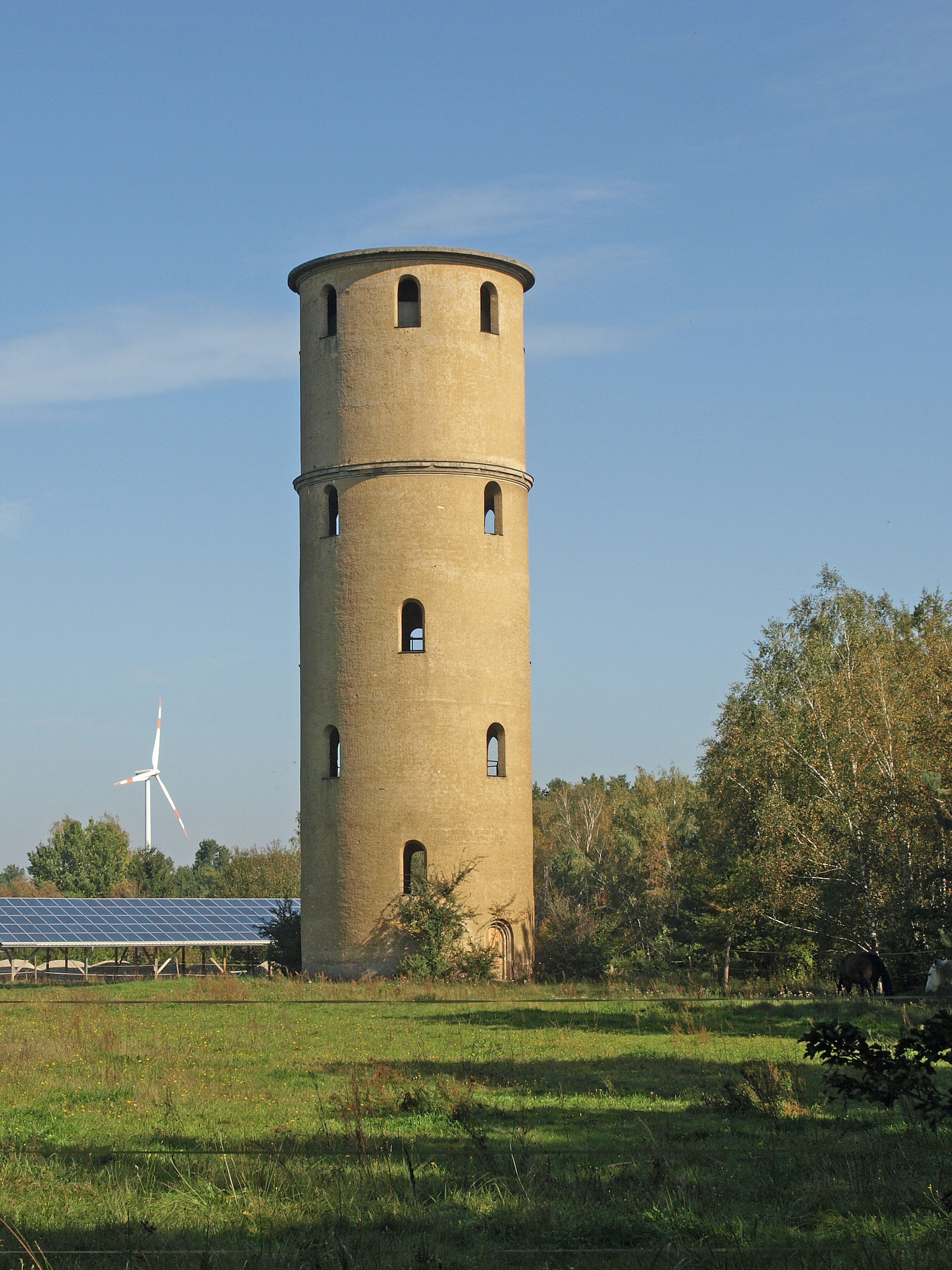 A tall, cylindrical tower with multiple windows, surrounded by a grassy field and trees. A wind turbine is visible in the background.
