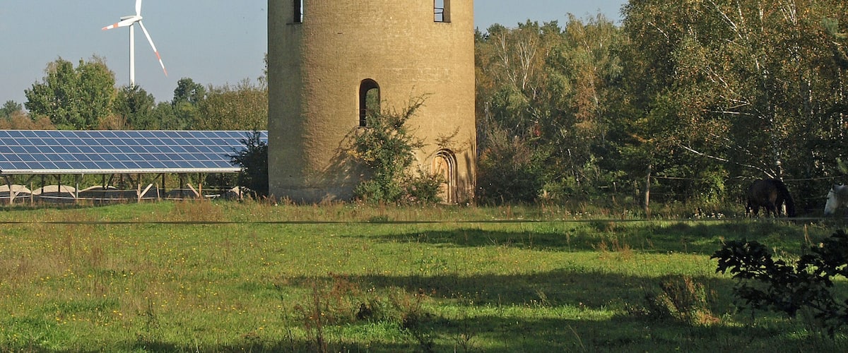 A tall, cylindrical tower with multiple windows, surrounded by a grassy field and trees. A wind turbine is visible in the background.