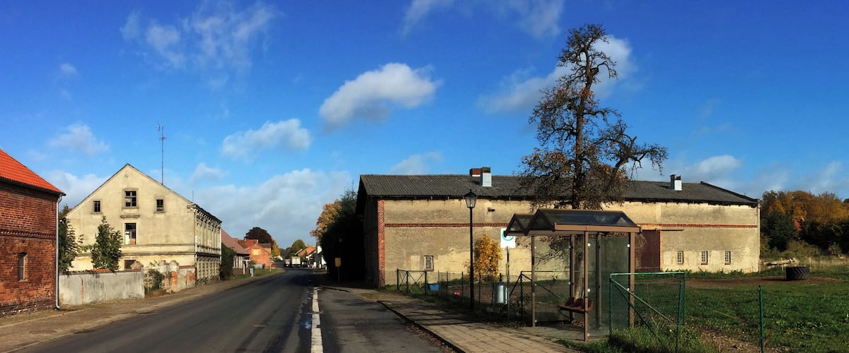 A rural street with old buildings, a green field, and a clear blue sky.