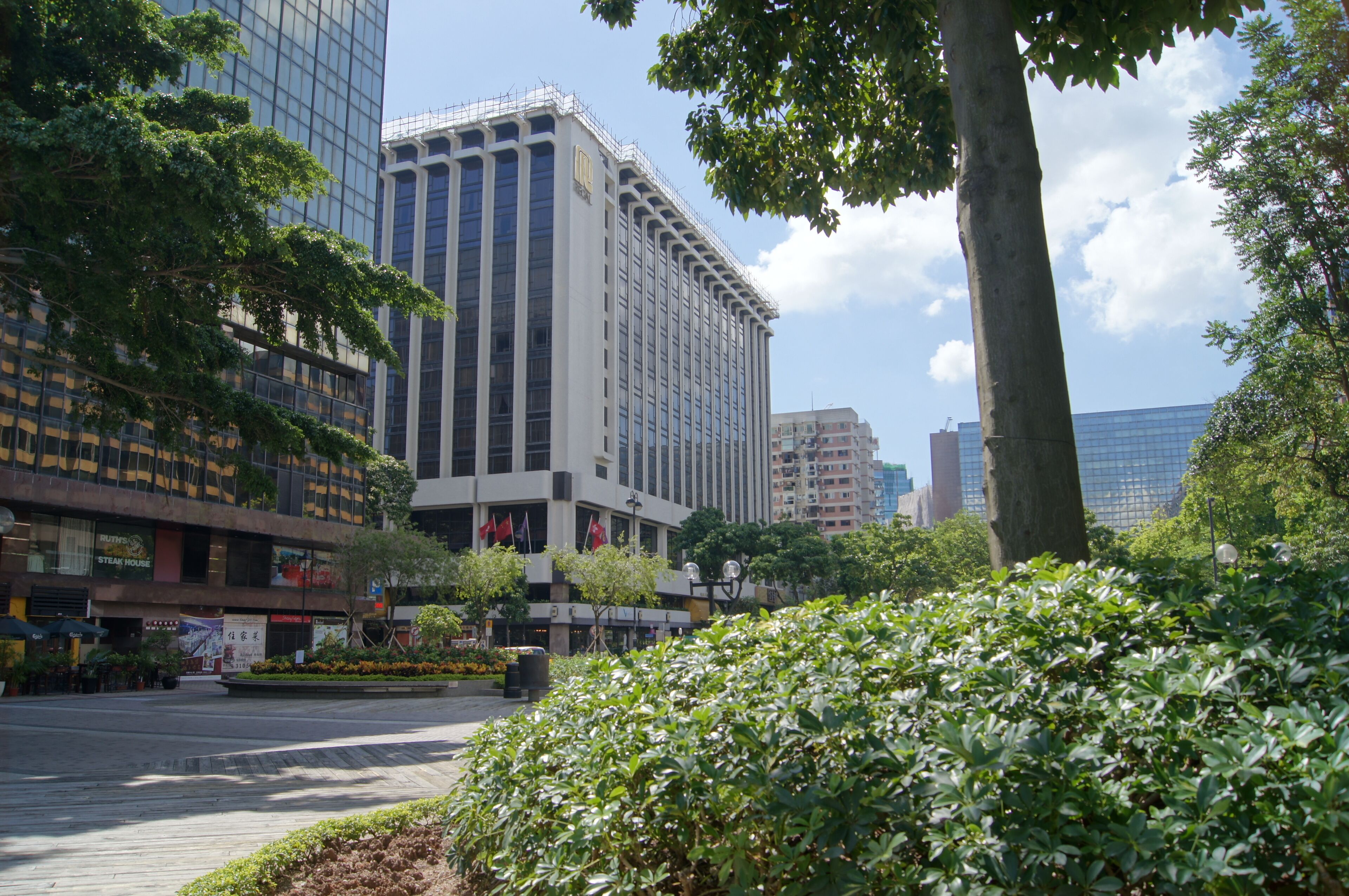 A cityscape with modern high-rise buildings, a central plaza with trees, and a clear sky.