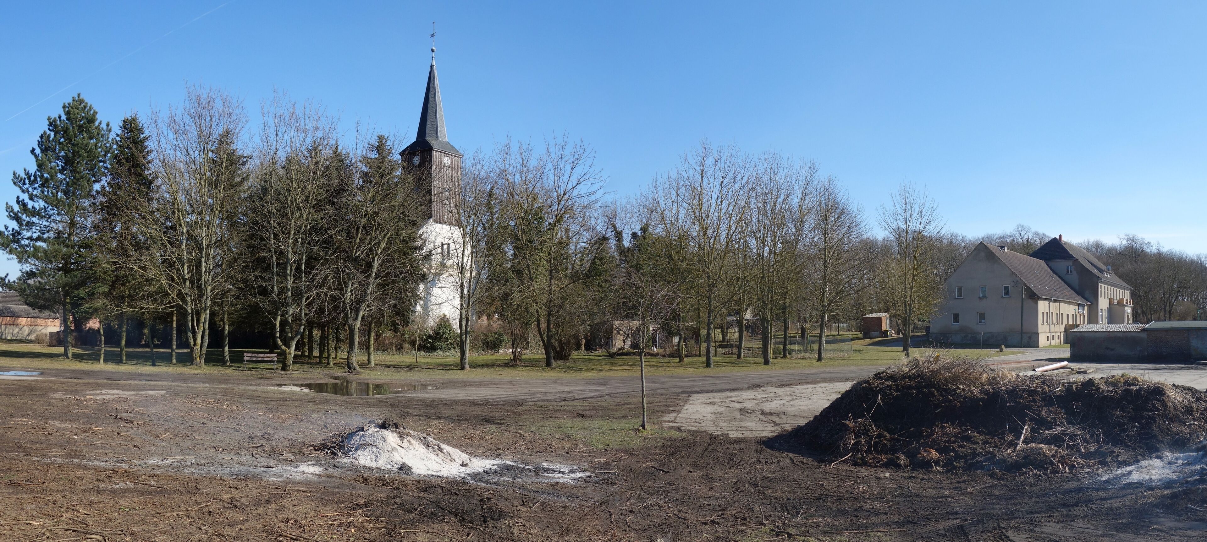 A church with a tall steeple, a large open field, and a building with a sloped roof.