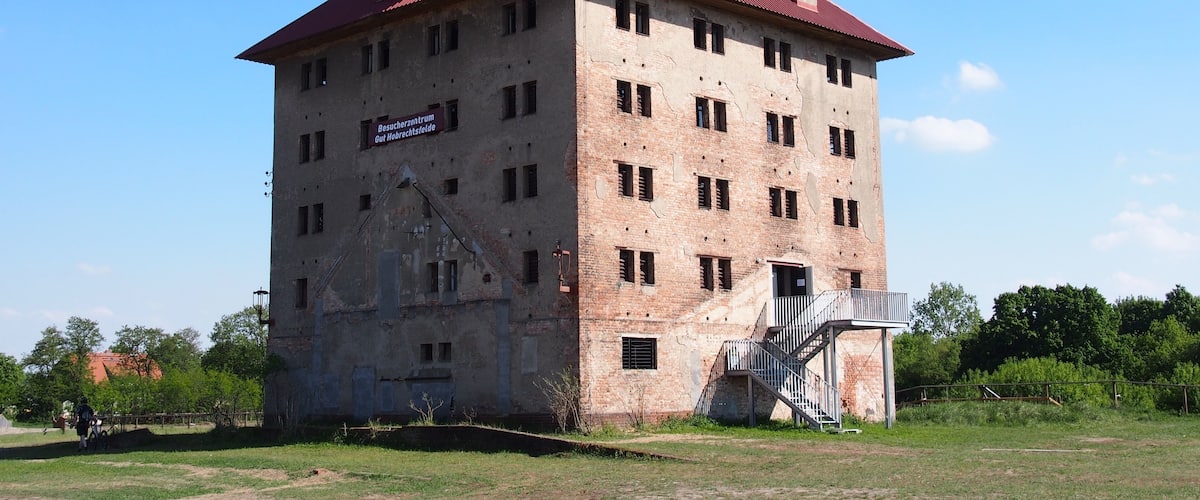 A large, multi-story brick building with a red roof and a sign that reads "Besucherzentrum Gut Mauerfeld."