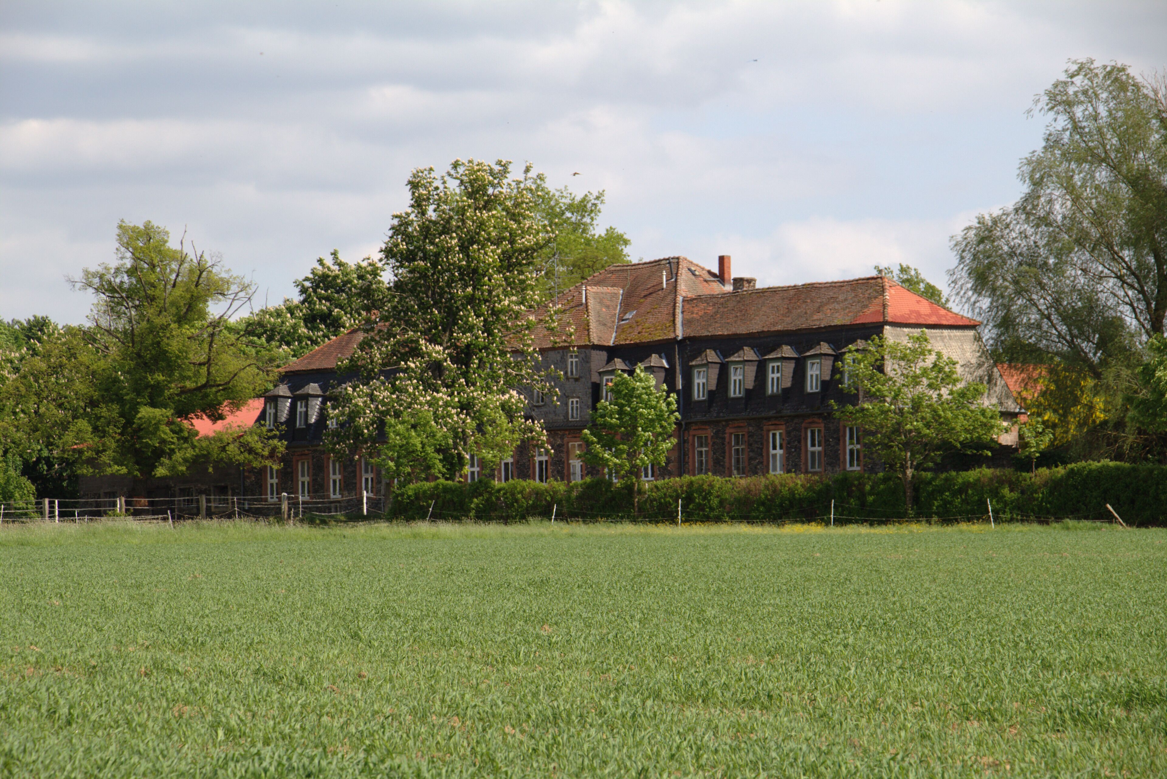 A large, historic building with a red roof, surrounded by a well-maintained grassy field and trees.