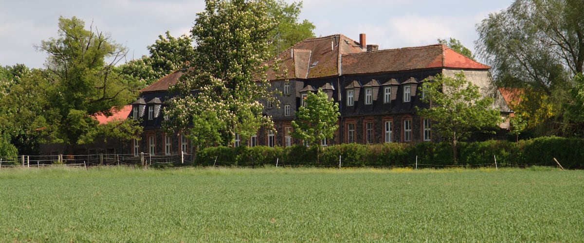 A large, historic building with a red roof, surrounded by a well-maintained grassy field and trees.