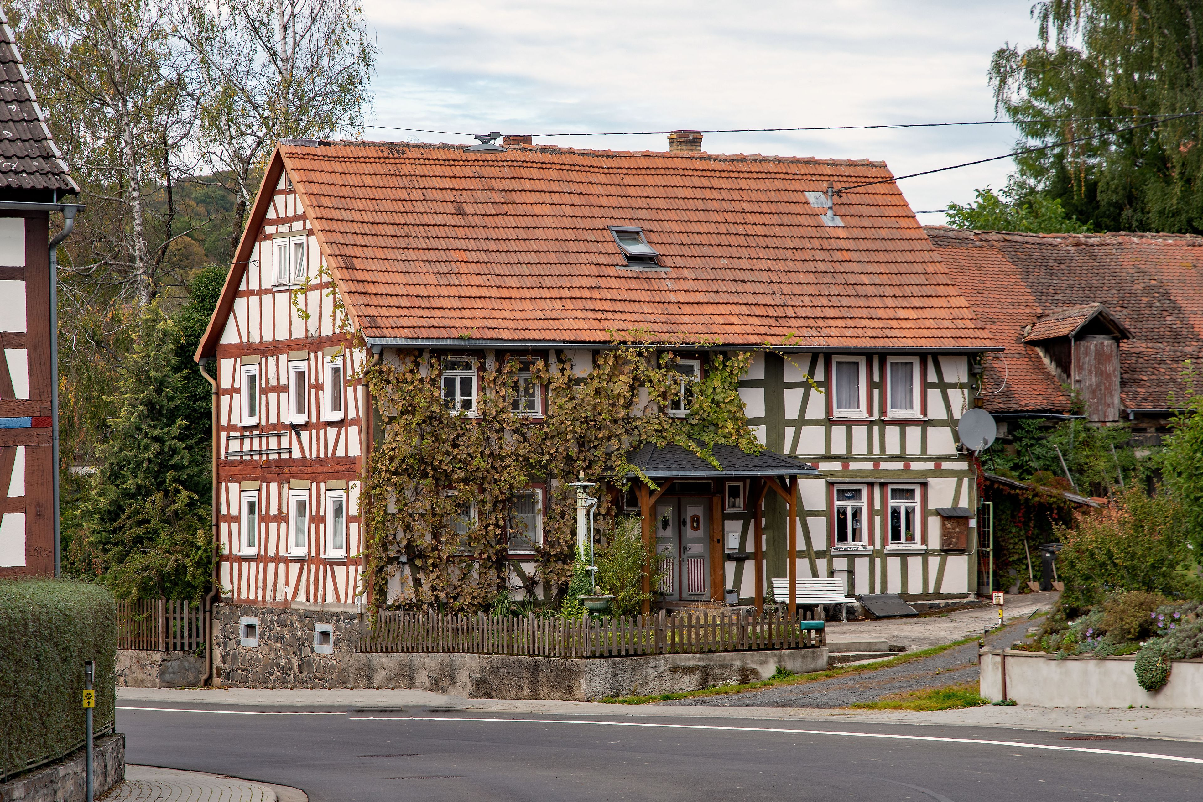 A traditional half-timbered house with a red-tiled roof, surrounded by greenery and a stone wall.
