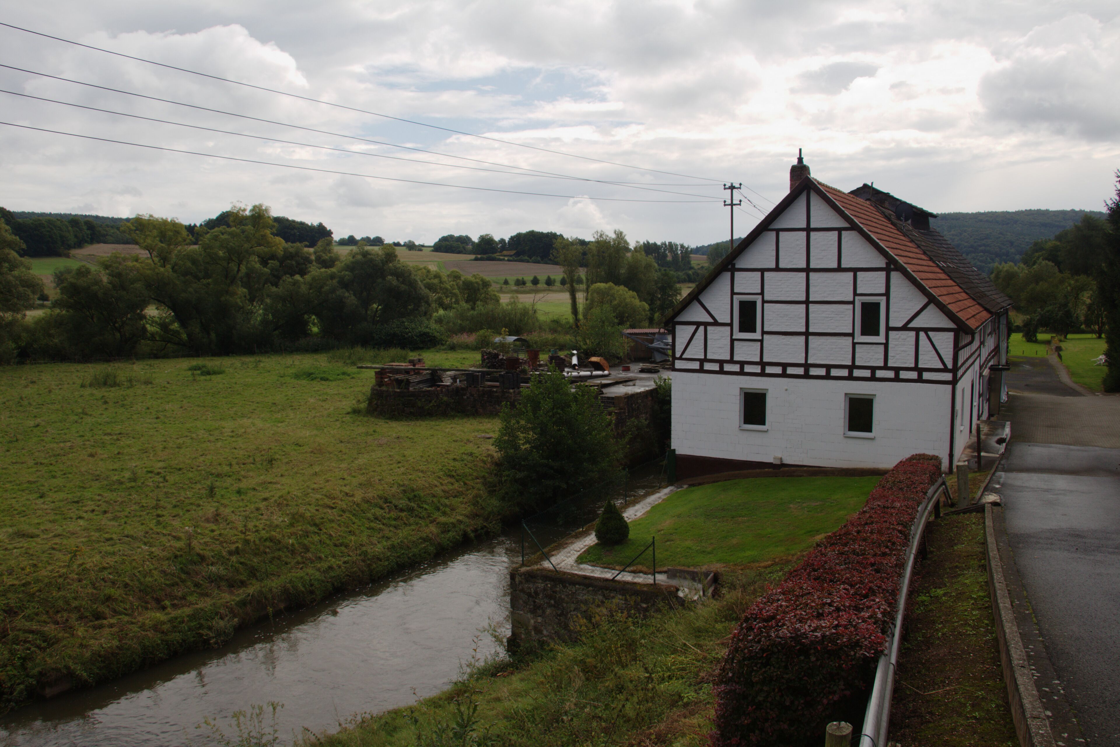 A white half-timbered house with a red roof, a narrow canal, and a green landscape with trees and fields.