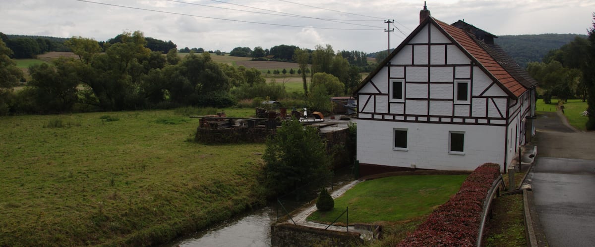 A white half-timbered house with a red roof, a narrow canal, and a green landscape with trees and fields.