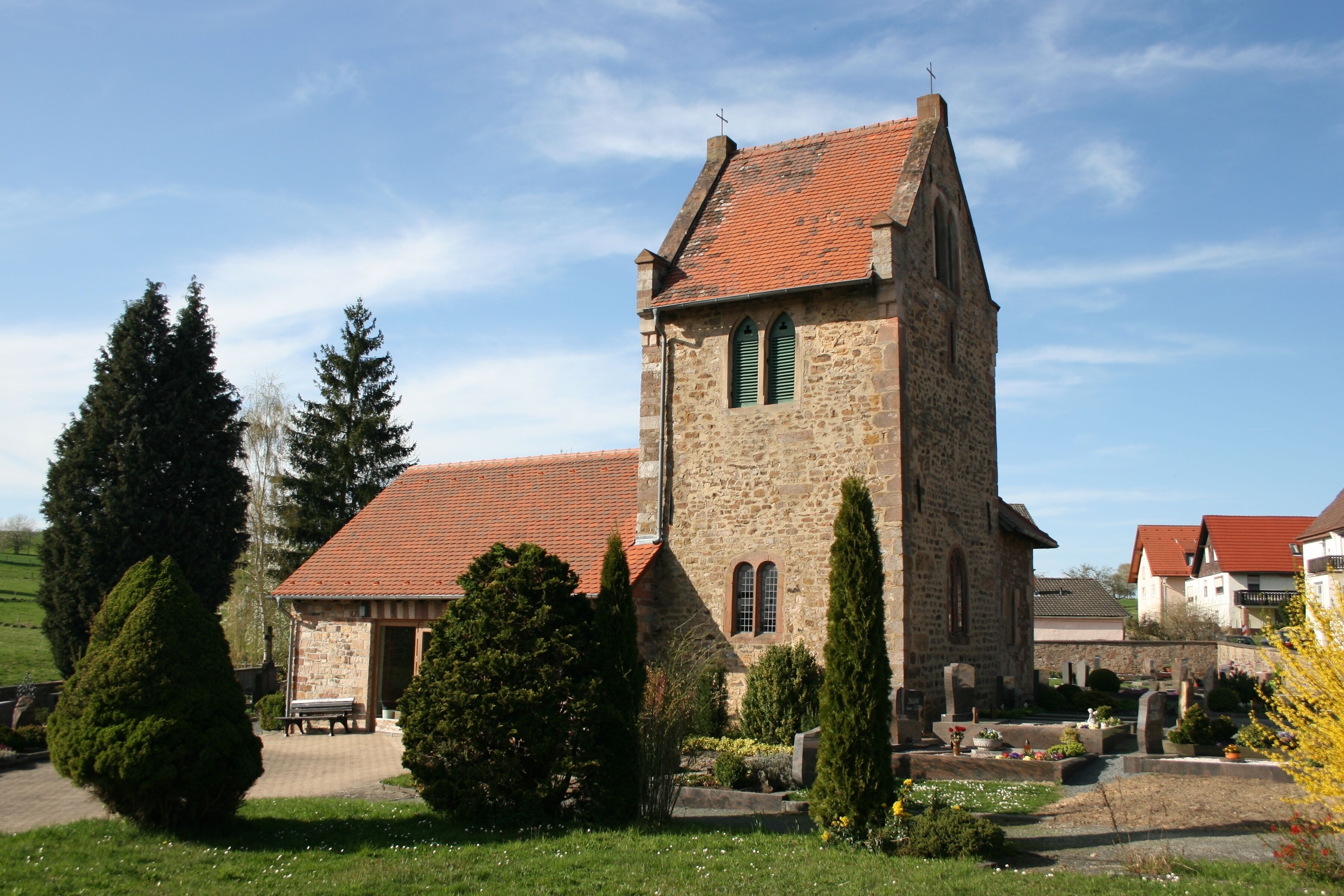 A stone church with a red-tiled roof, surrounded by greenery and a cemetery.