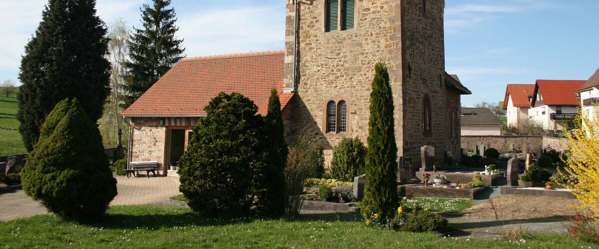 A stone church with a red-tiled roof, surrounded by greenery and a cemetery.