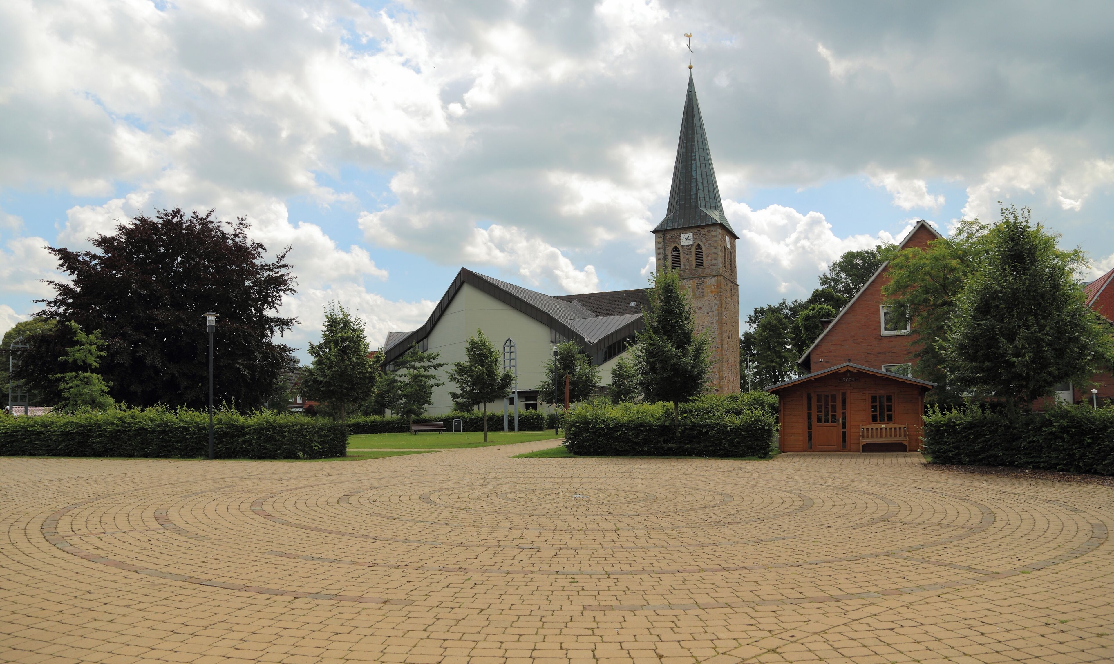 A church with a tall steeple and a clock tower, surrounded by trees and a paved courtyard.