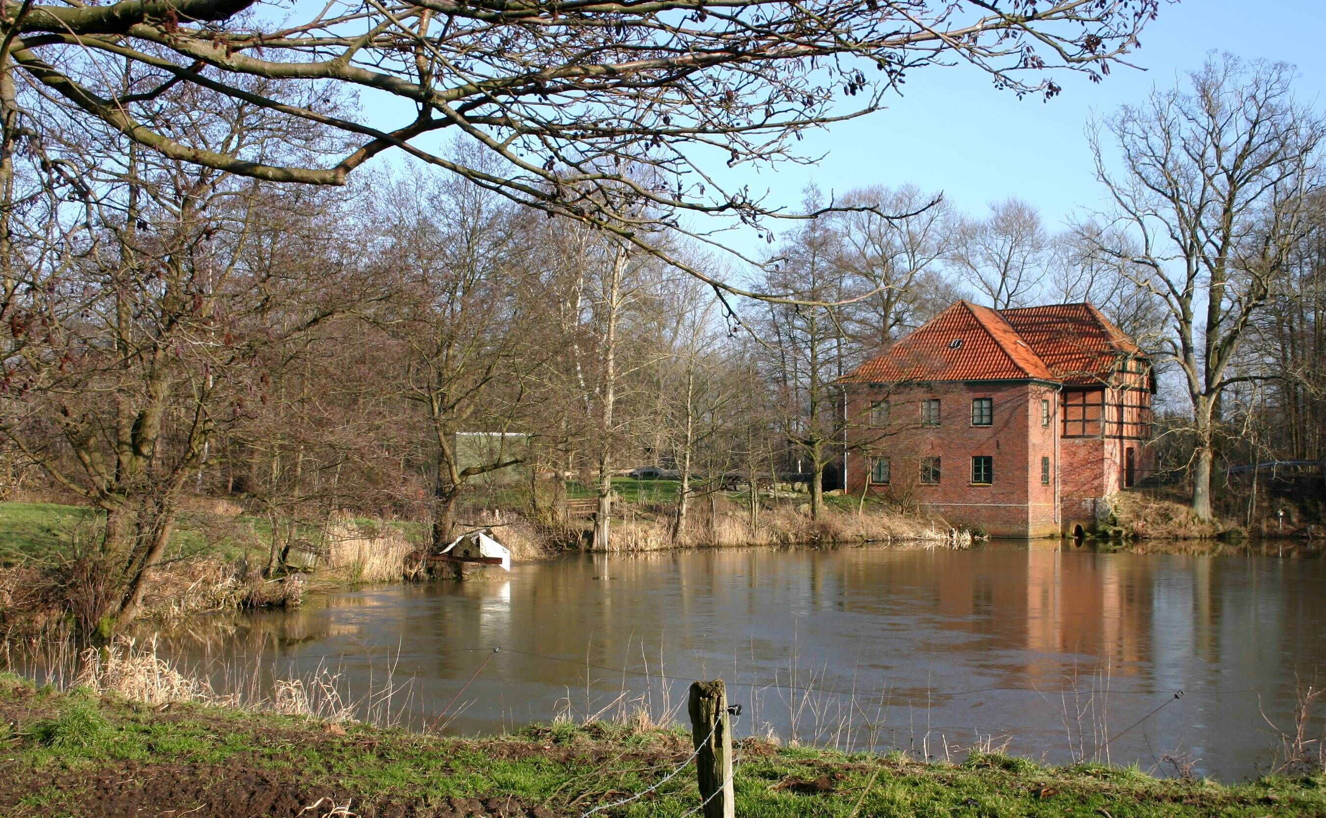 A red brick house by a calm river with trees in the background.