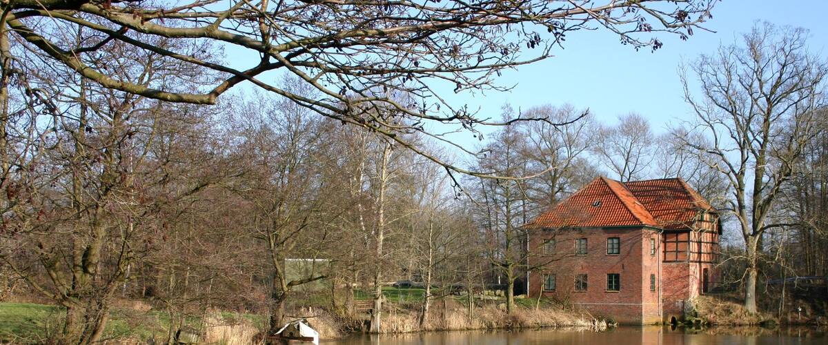 A red brick house by a calm river with trees in the background.