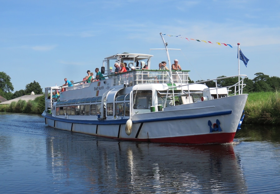 Un barco de recreo con pasajeros, una bandera y una vía fluvial tranquila.