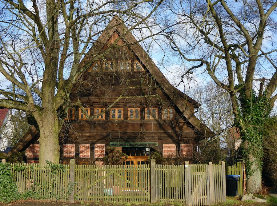A traditional wooden house with a steeply pitched roof, surrounded by trees and a wooden fence.