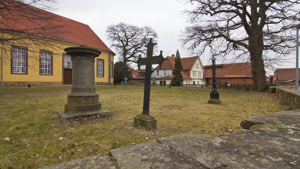 A cemetery with a yellow building, stone crosses, and a large tree.