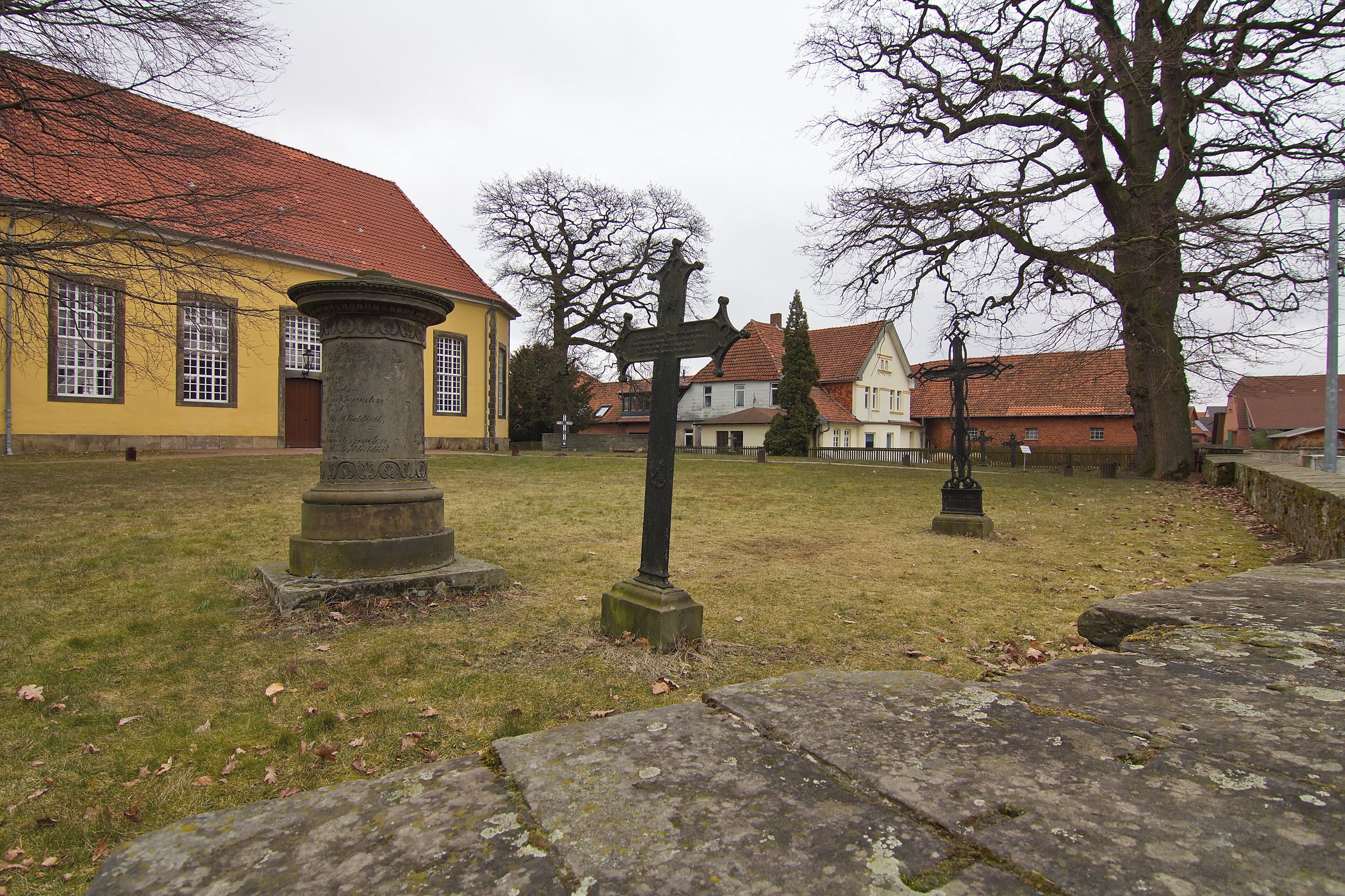 A cemetery with a yellow building, stone crosses, and a large tree.