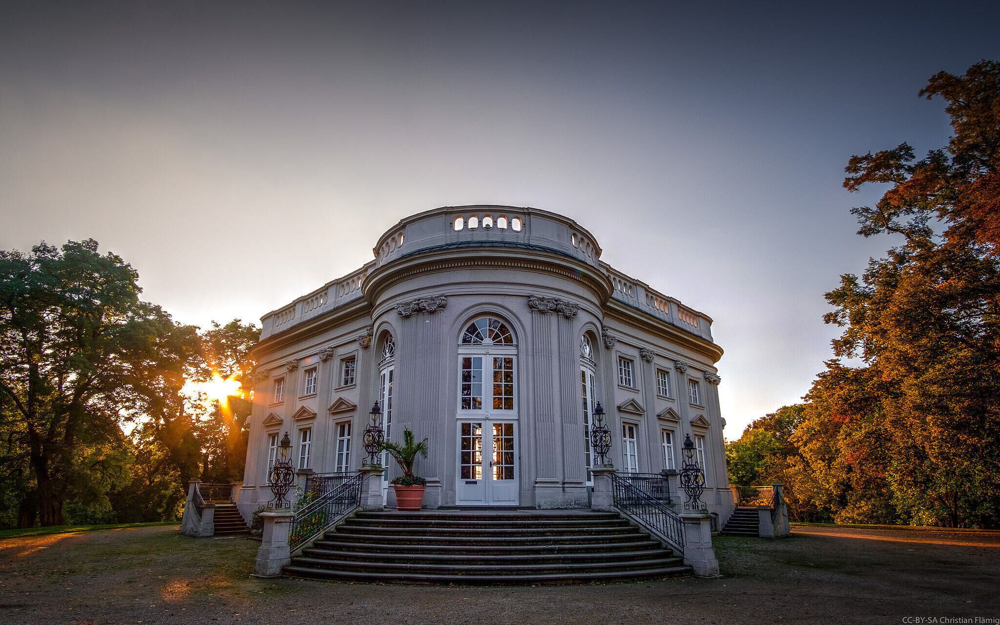 A classical building with a grand entrance, surrounded by trees and a sunset in the background.