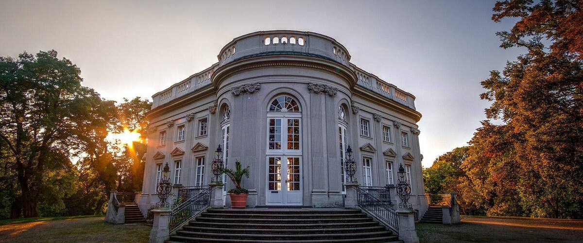 A classical building with a grand entrance, surrounded by trees and a sunset in the background.