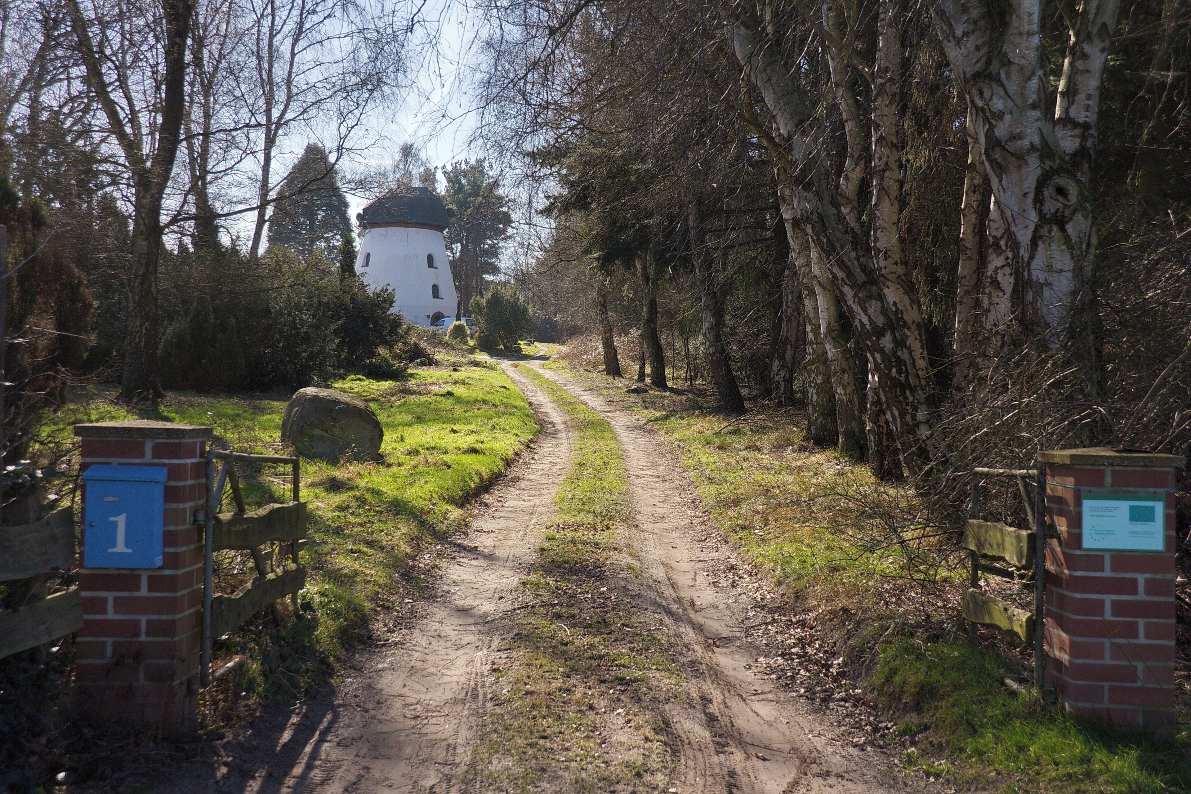 A dirt path leading to a white building with a dome roof, surrounded by trees and a wooden gate with a blue sign.