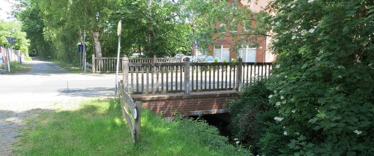A brick bridge over a small stream, surrounded by lush greenery and a wooden fence.
