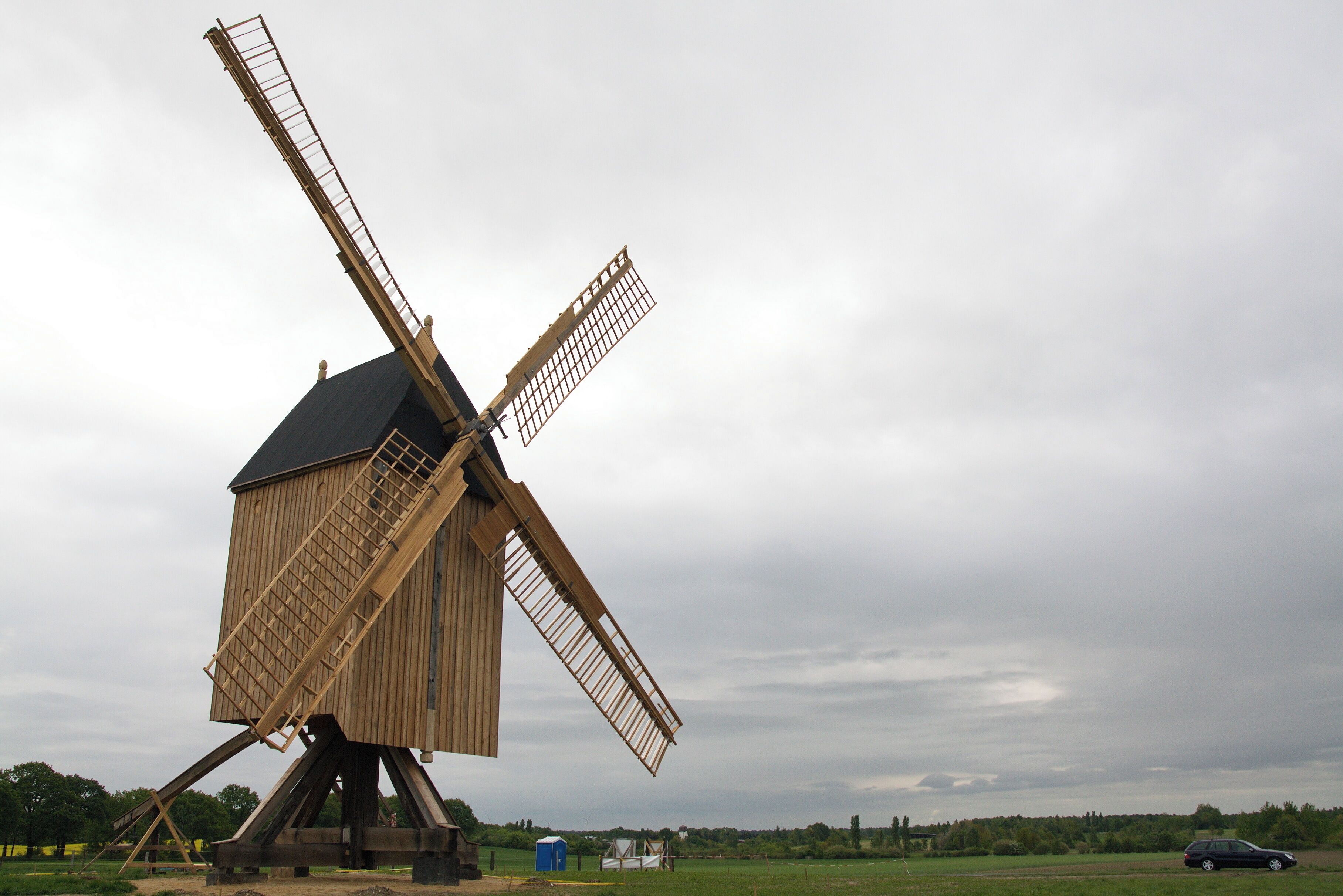 A large wooden windmill with a tilted structure, set against a cloudy sky.