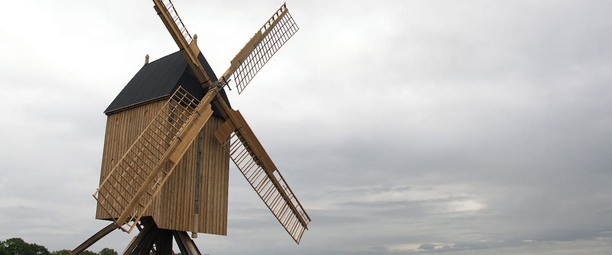 A large wooden windmill with a tilted structure, set against a cloudy sky.