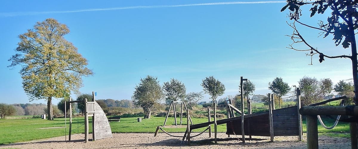 A playground with swings and a slide, surrounded by trees and open grassy areas.