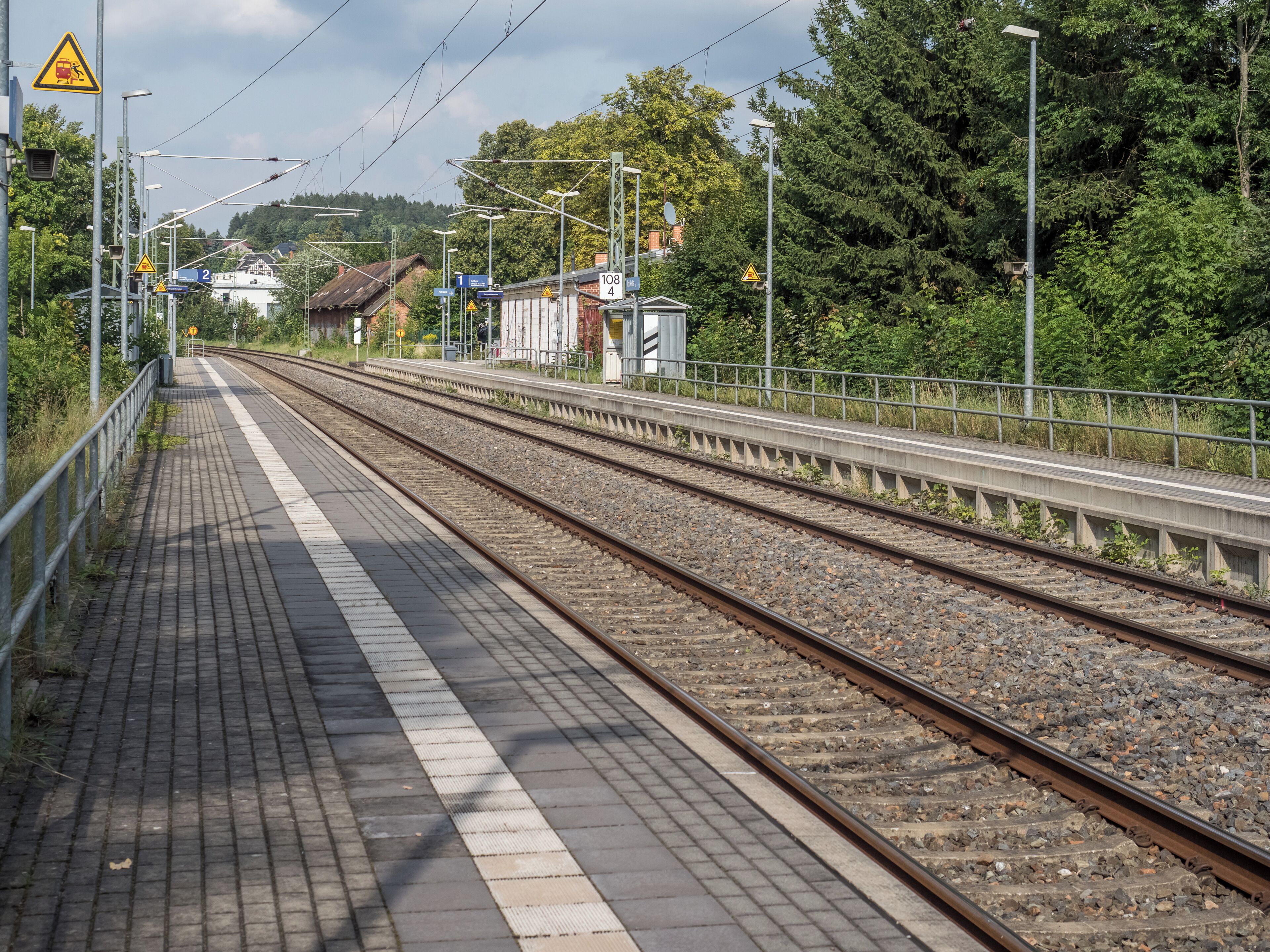 A railway station platform with multiple tracks, overhead wires, and surrounding greenery.
