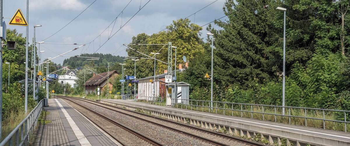 A railway station platform with multiple tracks, overhead wires, and surrounding greenery.