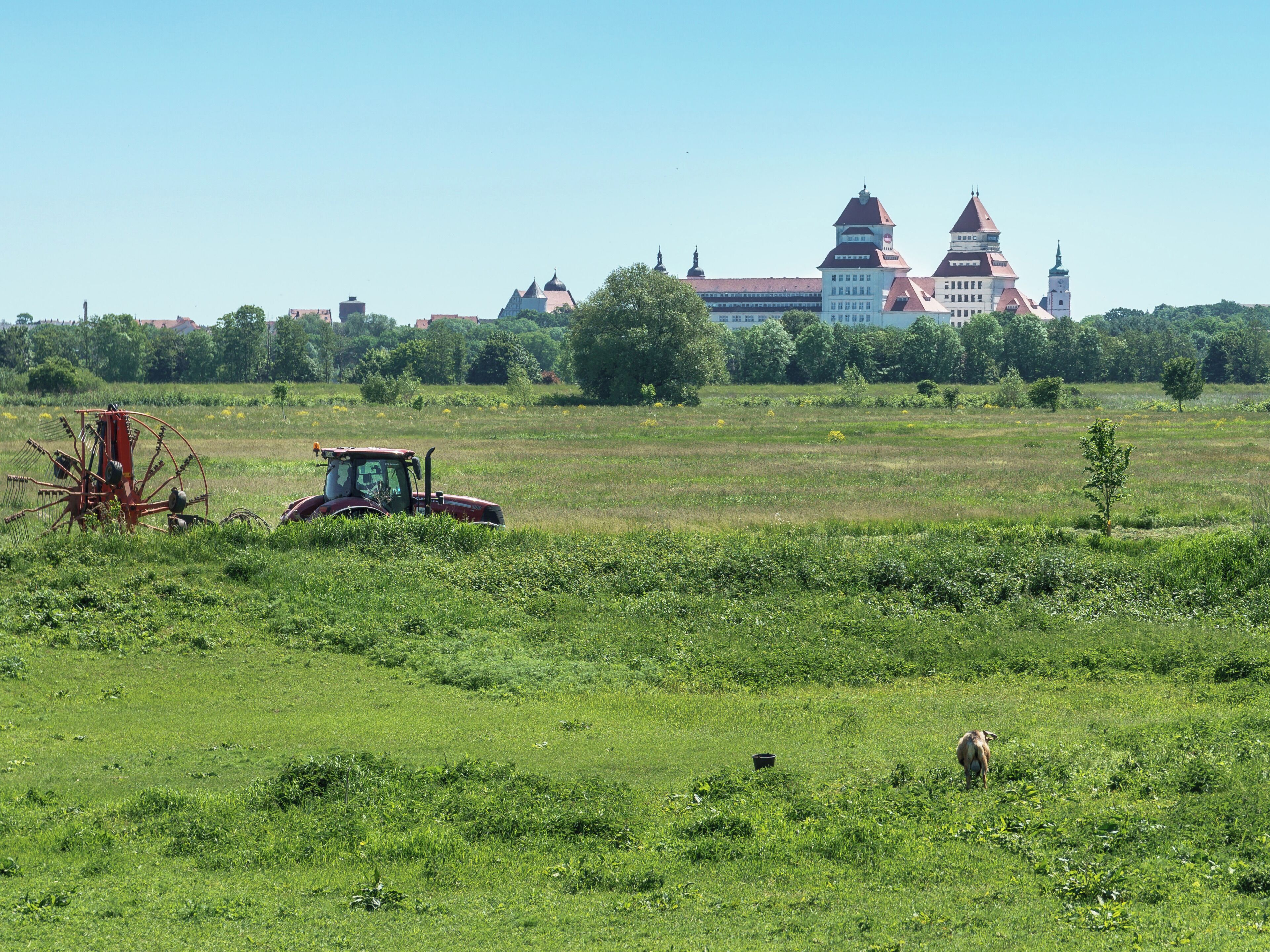 A green field with a tractor and a large building with a red roof in the background.